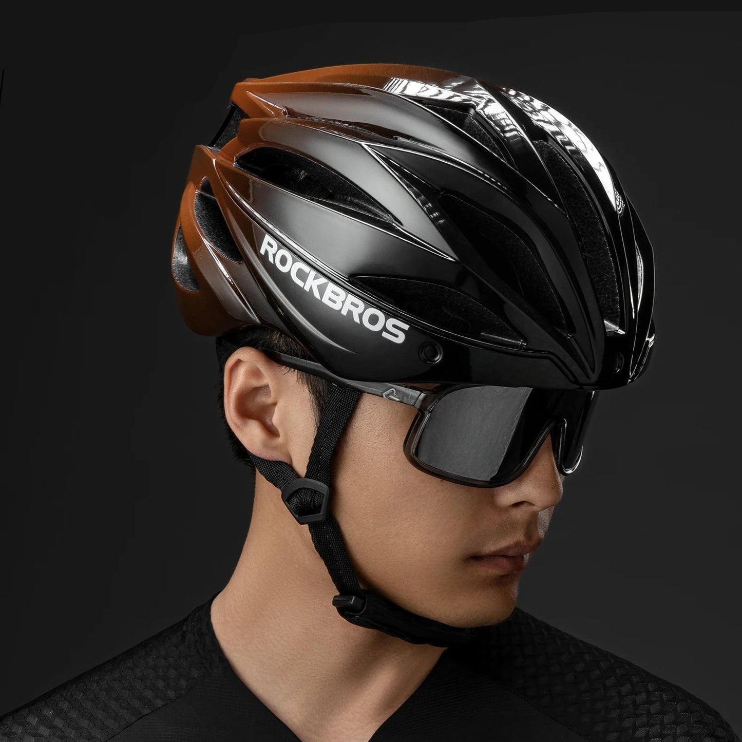 A man in cycling gear wearing a glossy black and brown Rockbros cycling helmet with matching black sunglasses, shown in profile against a dark studio background. The Rockbros logo is visible in the top-left corner.